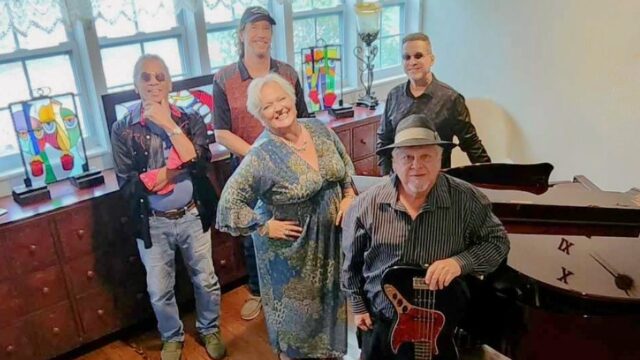 Five people stand indoors near a grand piano, with two colorful stained glass lamps on a cabinet behind them. One person holds an electric guitar.