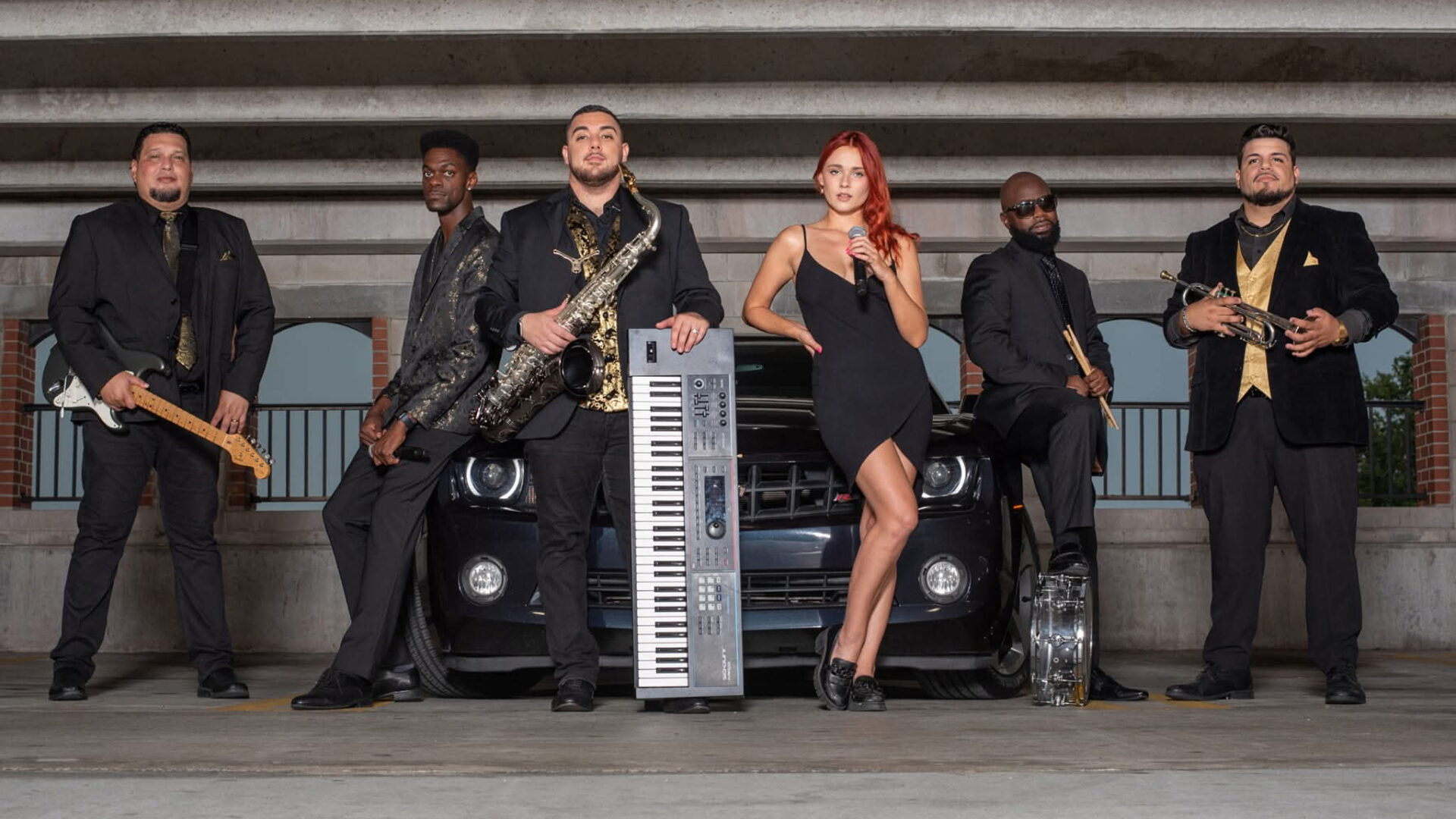 Six musicians pose in front of a black car in a parking garage, holding instruments including a saxophone, keyboard, guitar, and trumpet.