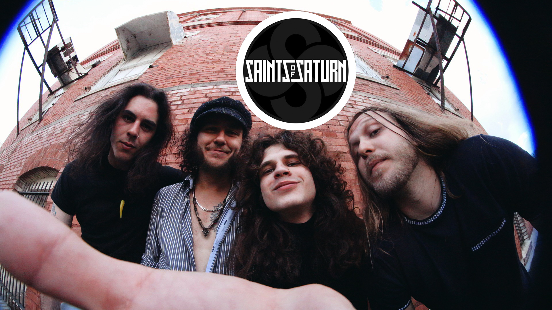 Four men with long hair stand in front of a brick building, smiling at the camera; a 