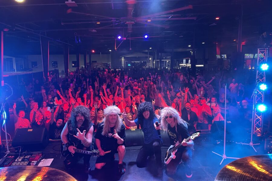 Four rock musicians with long hair pose on stage with guitars in front of a large, enthusiastic crowd at an indoor concert venue, illuminated by colorful stage lights.