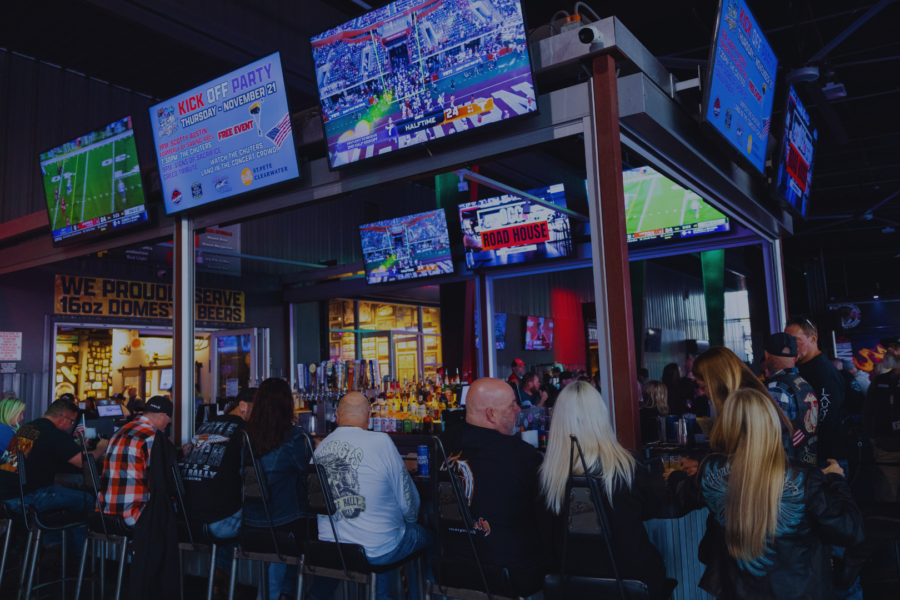 People sit at a bar watching multiple large TVs displaying sports games in a dimly lit sports bar setting.