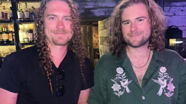Two men with long curly hair stand side by side in a bar, one wearing a black shirt, the other a green shirt with floral designs. Shelves with bottles are visible in the background.