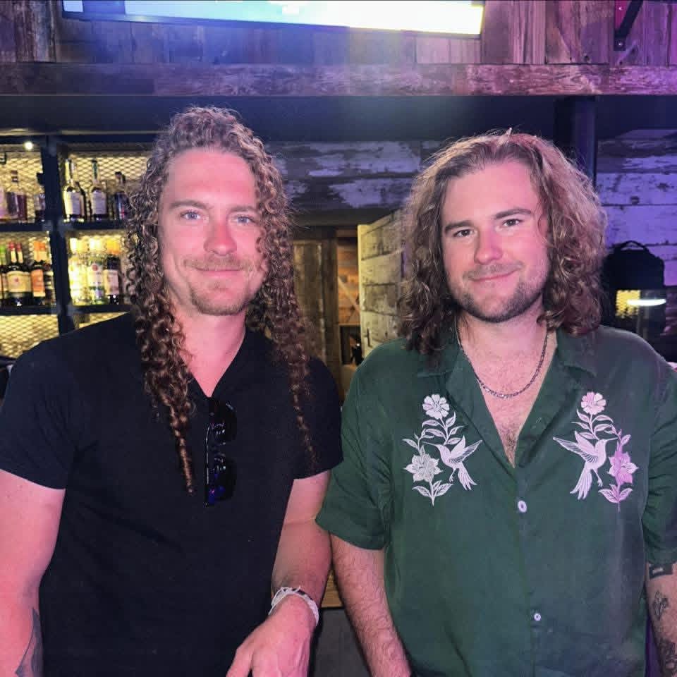 Two men with long curly hair stand side by side in a bar, one wearing a black shirt, the other a green shirt with floral designs. Shelves with bottles are visible in the background.