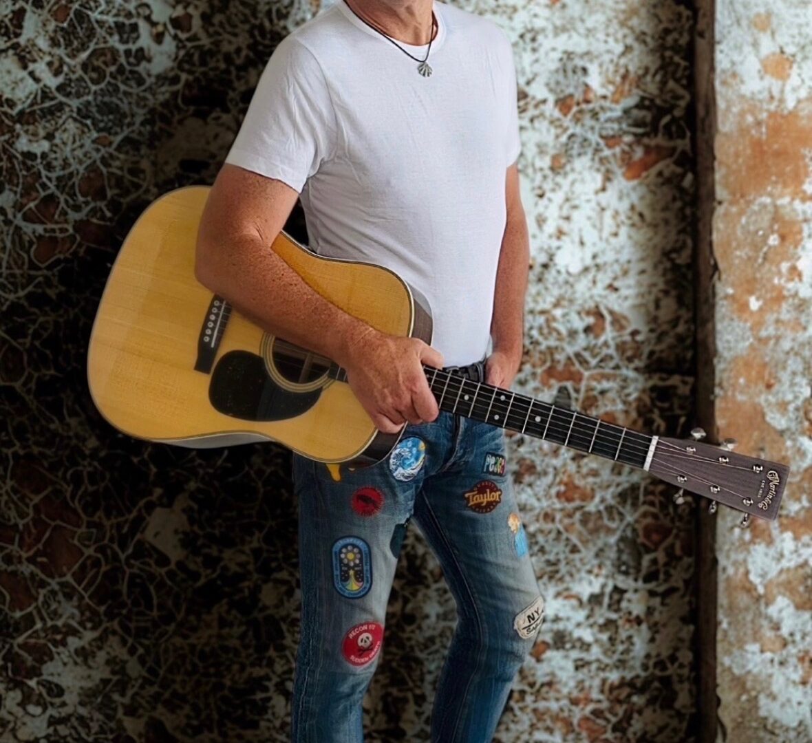 A man in a white t-shirt and patched jeans standing while holding an acoustic guitar.