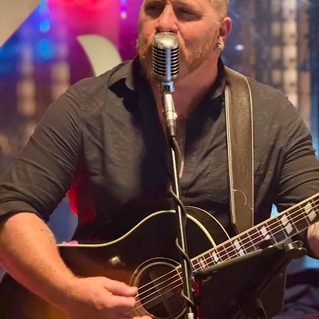 A man with a beard singing into a vintage-style microphone while playing an acoustic guitar.